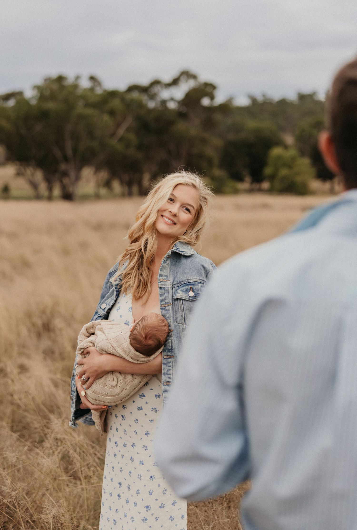 A smiling blonde woman holds a newborn baby in a golden wheat field, wearing a denim jacket and floral dress.