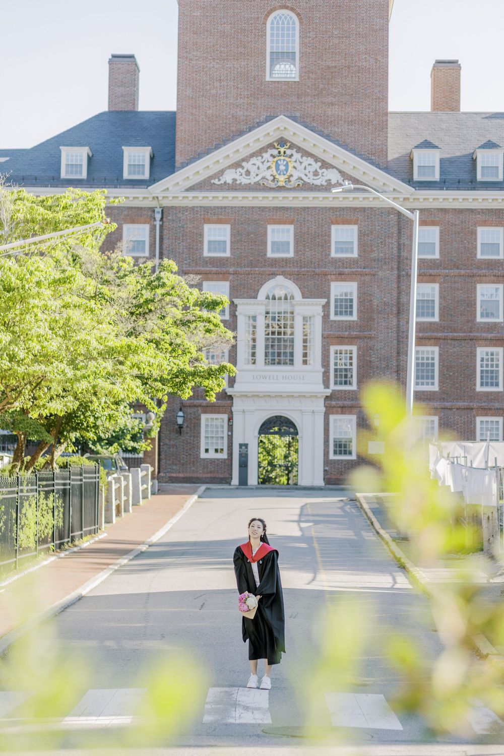 Harvard Graduate in cap and gown stands before a grand brick university building in Harvard on a sunny day.