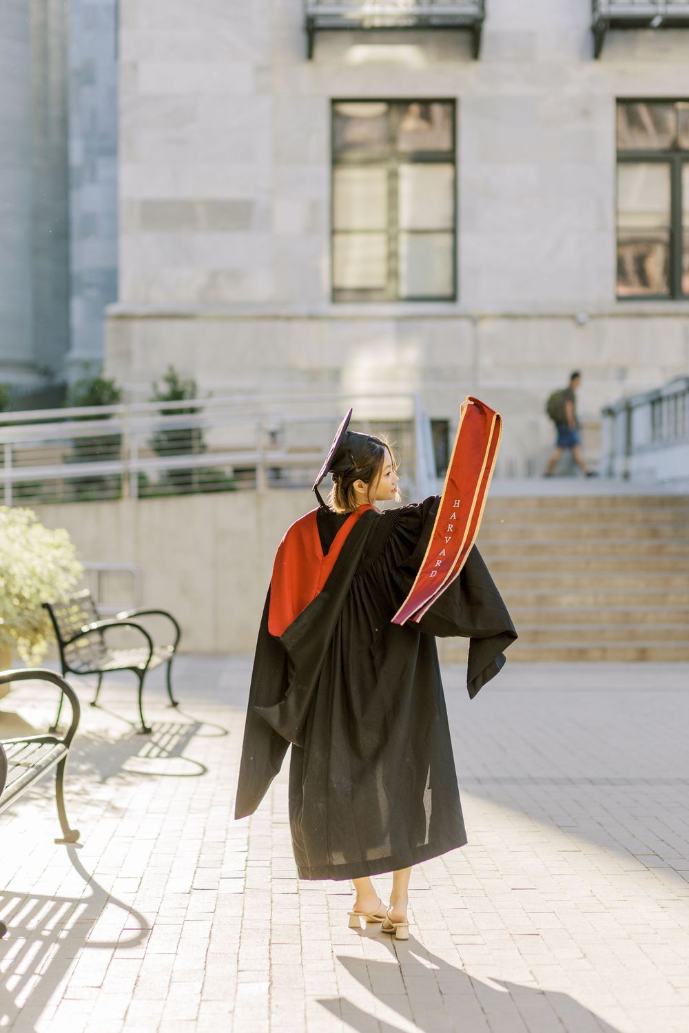 Harvard Graduate in black cap and gown holds diploma outdoors on campus plaza, facing away from camera.