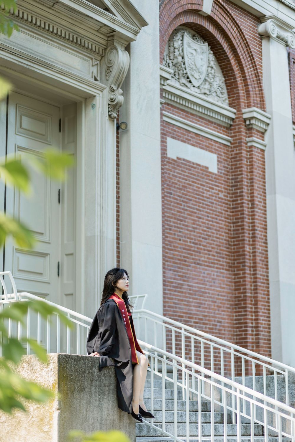 A young woman in Harvard graduation gown and red stole poses on steps of a classic brick and stone university building.