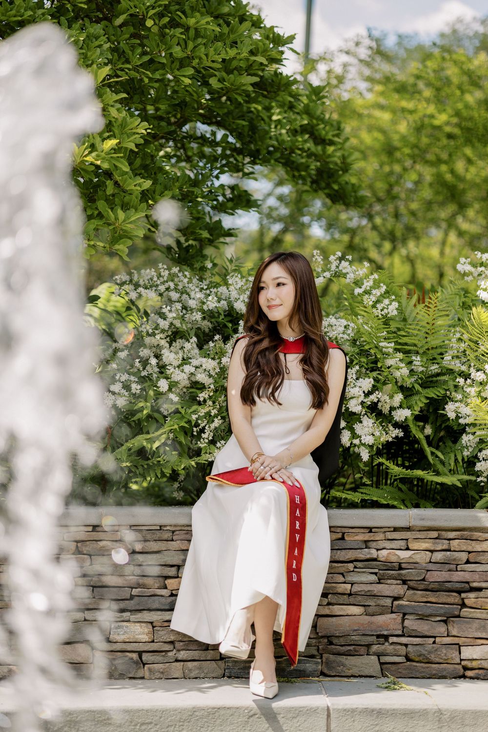 Young Asian woman in white and red dress sits on stone wall surrounded by lush green foliage and white flowers at Harvard for graduation