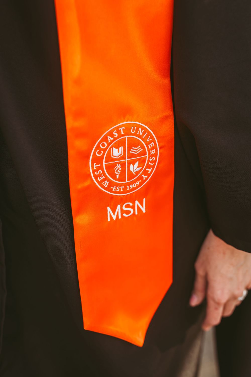 Close-up of orange West Coast University MSN graduation stole on black academic gown.