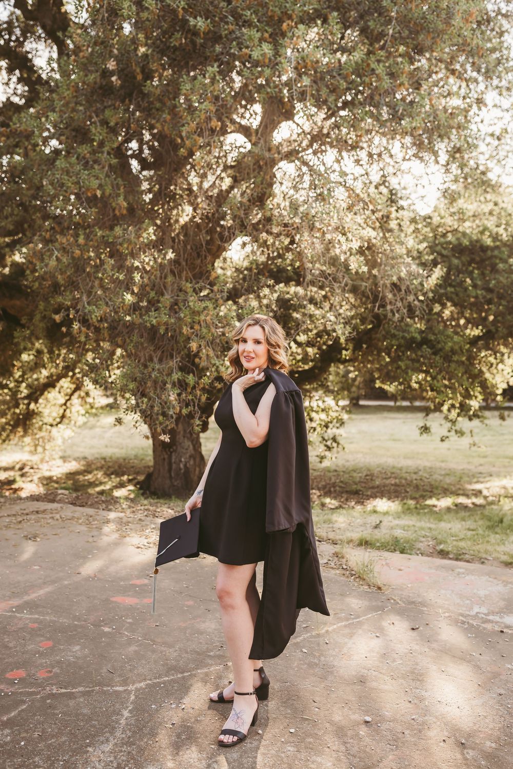 Smiling graduate in black dress and gown holds cap outdoors under large oak tree in golden light.