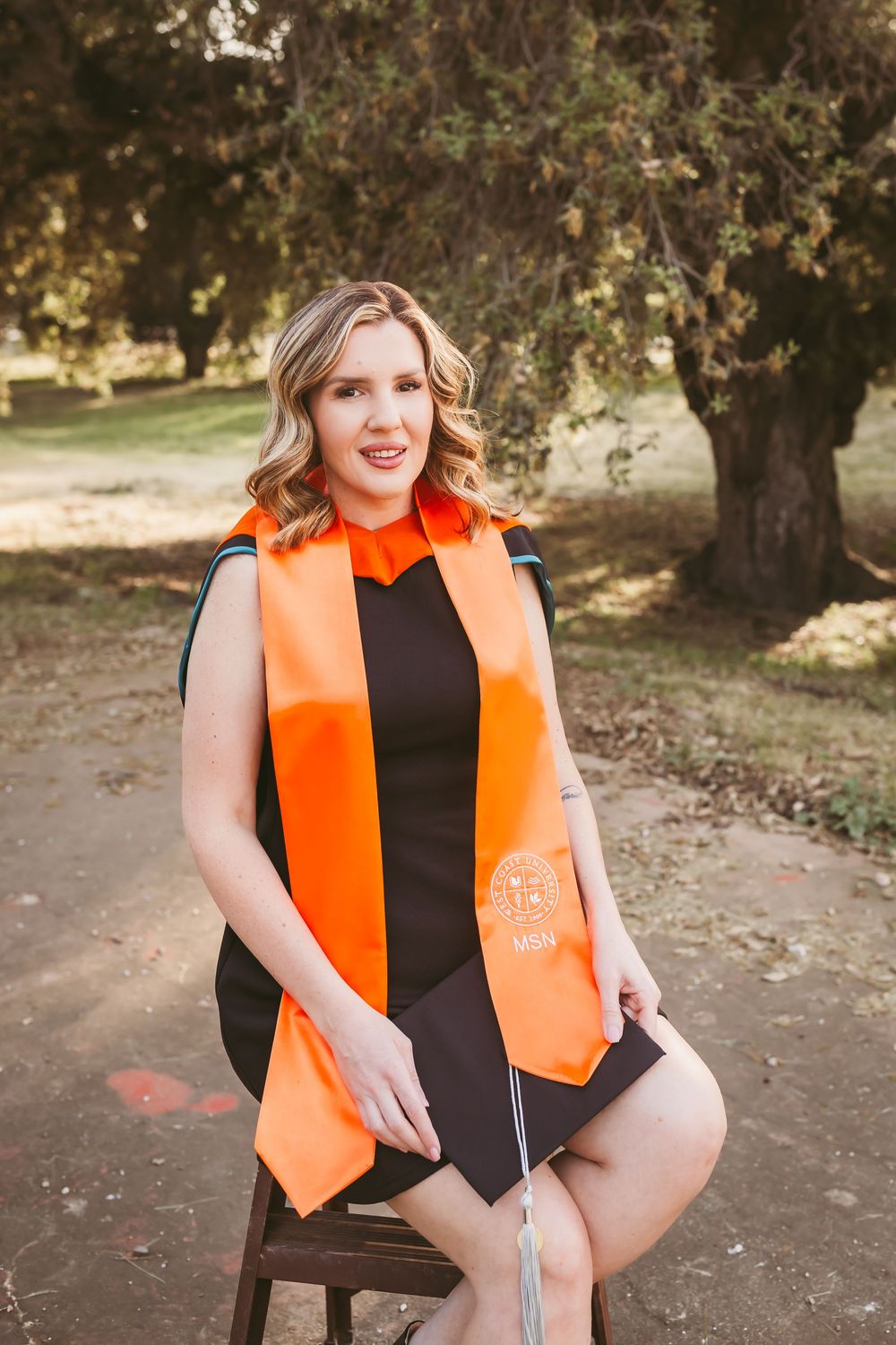 Young woman in black dress and orange graduation stole sits outdoors holding cap, smiling for graduation portrait.
