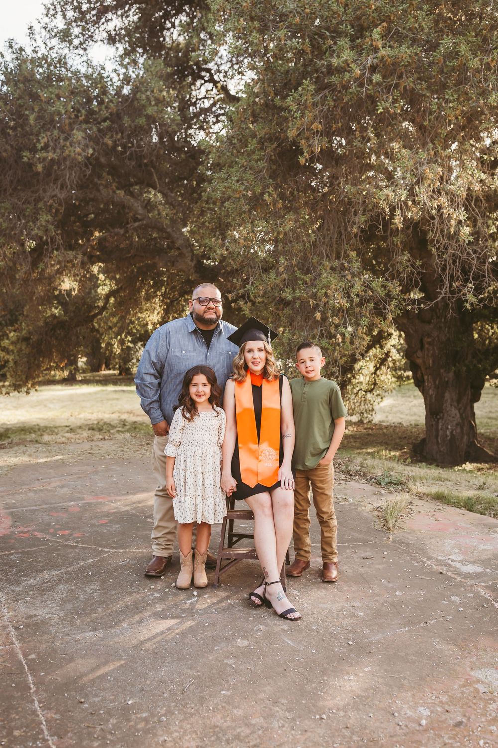 Family of four poses outdoors under oak trees, mom wearing orange graduation cap and sash celebrating her achievement near yucaipa ca