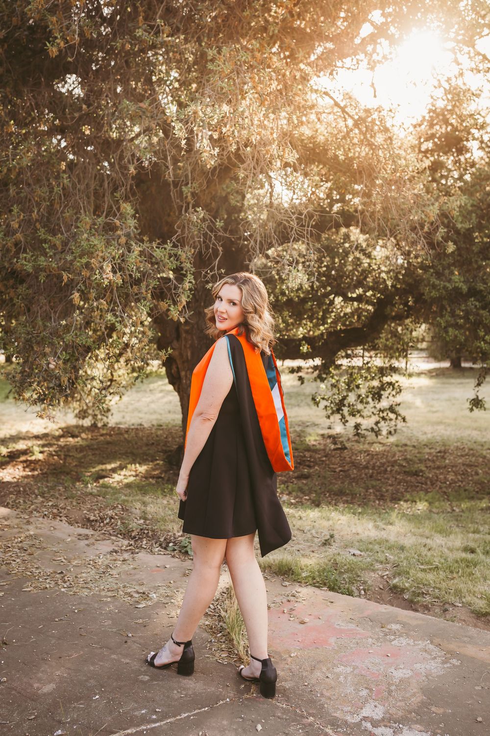 Young woman in black dress with orange sash poses outdoors among trees in golden sunlight.