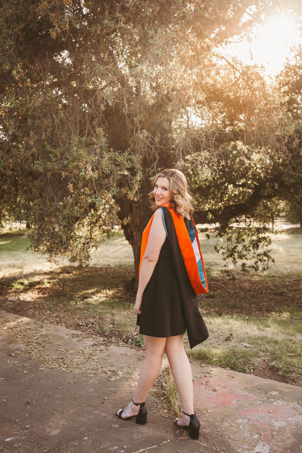 A smiling woman in a black dress and orange graduation sash poses outdoors under trees in golden sunlight.