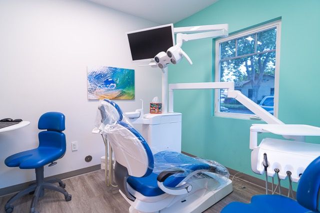 Modern dental exam room with blue chair, monitor, teal walls, and dental equipment in a clean, bright office setting.