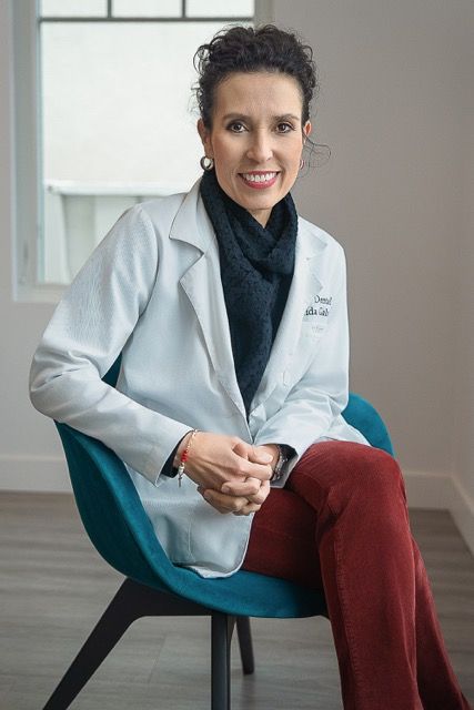 Female doctor in white lab coat and red pants sitting in teal chair, smiling professionally in modern medical office.