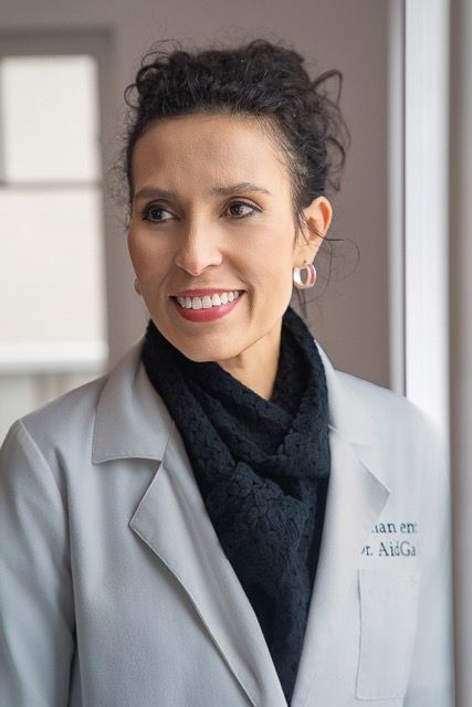 Female doctor in white lab coat smiling, wearing hoop earrings and black scarf, professional medical portrait.