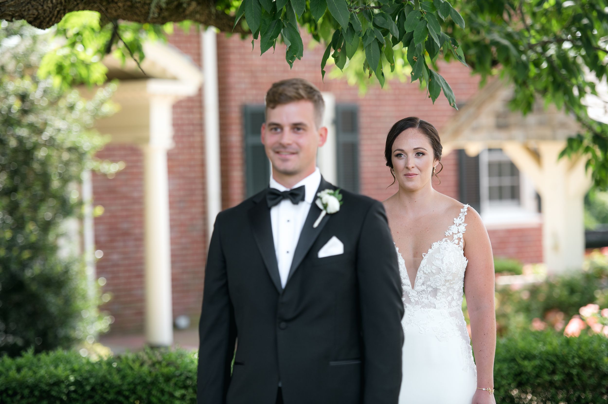 Bride in white gown peers around lush green bushes at groom in tuxedo during romantic first look at brick venue.