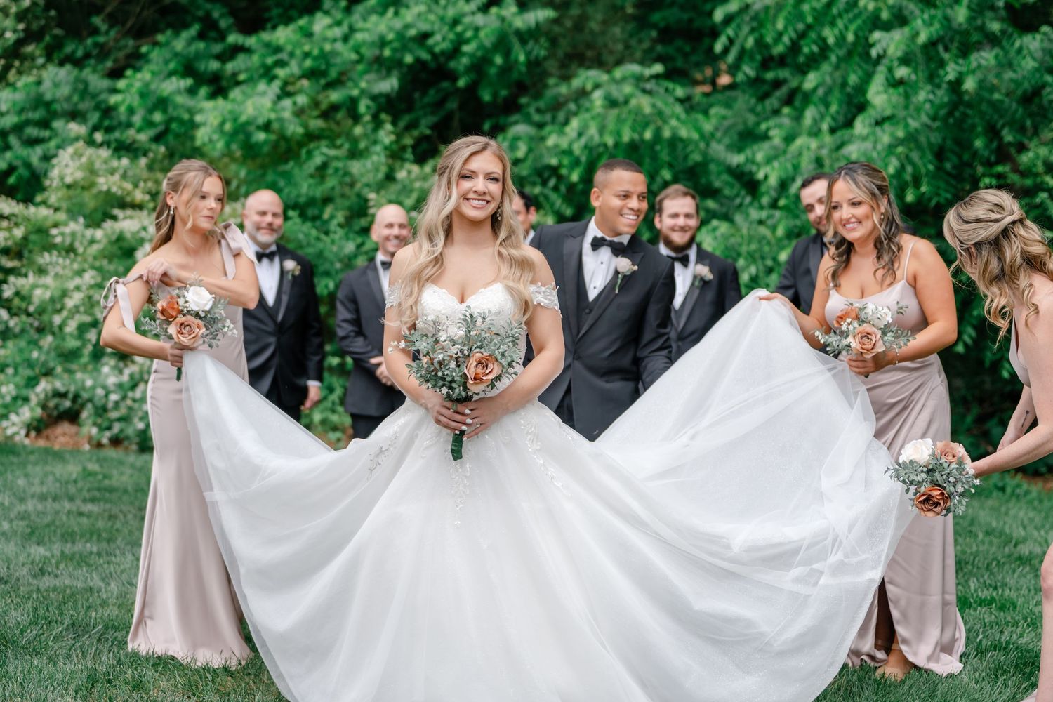 Bride in a white ball gown holds a bouquet while bridesmaids and groomsmen gather behind her on a lush green lawn.