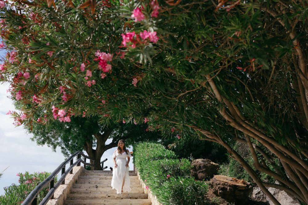 Woman in white dress walking up stone steps lined with blooming pink oleander trees overlooking the sea.