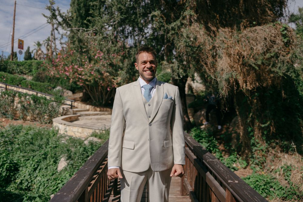Smiling groom in a light beige suit with blue tie standing on a wooden bridge surrounded by lush green trees.