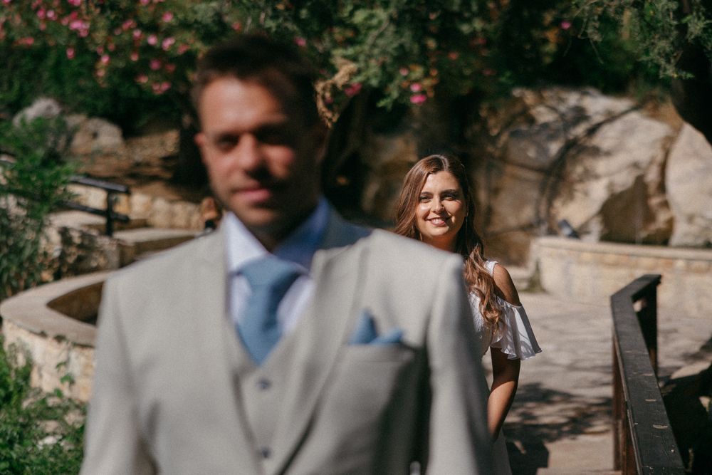 Bride smiling at groom in grey suit during outdoor wedding ceremony with lush greenery background.
