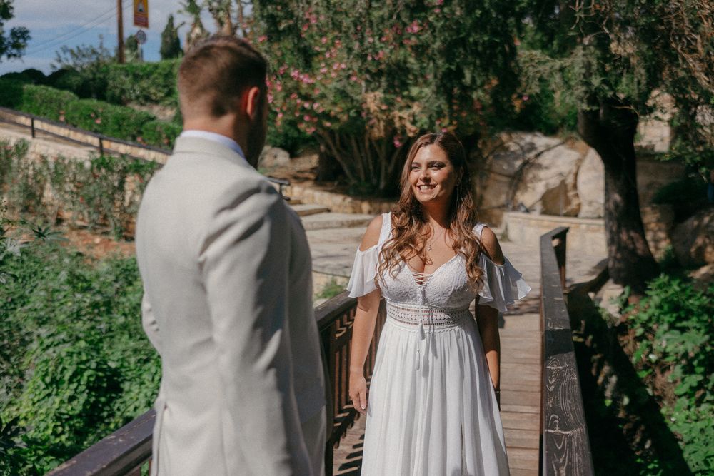 Bride in white boho dress smiles at groom in light suit on wooden bridge surrounded by lush greenery.