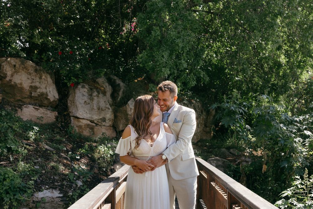 Couple sharing a romantic kiss on a wooden bridge surrounded by lush green trees during their outdoor wedding.