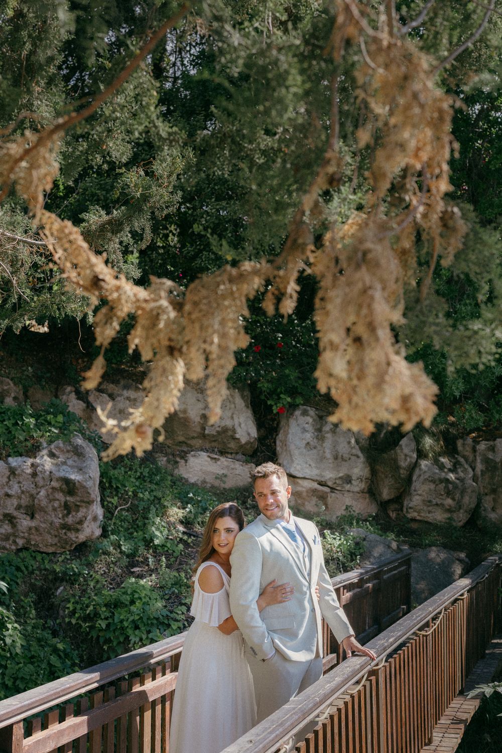 Bride and groom posing on a wooden bridge surrounded by lush greenery and rocky landscape on their wedding day.