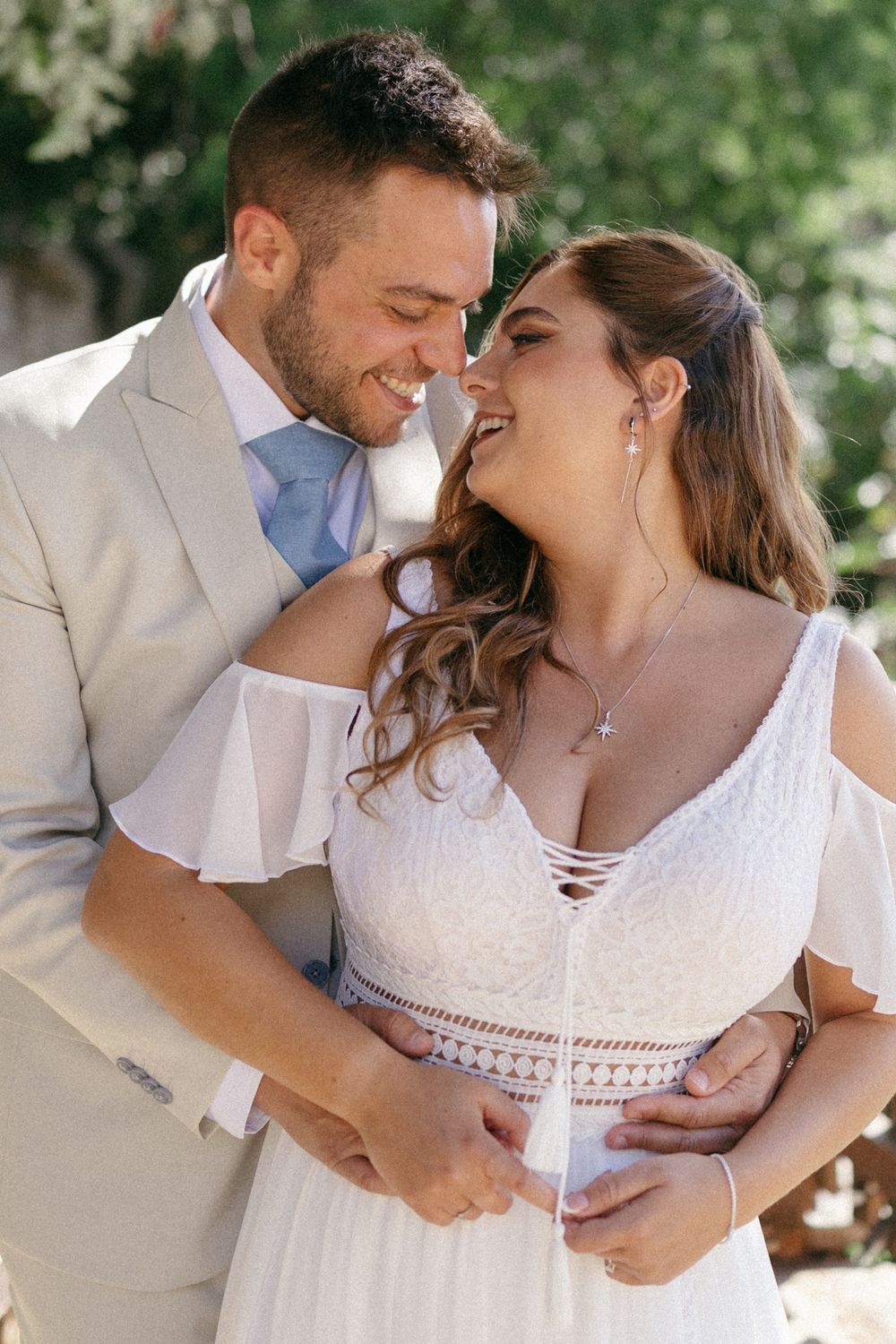 A smiling bride in white dress and groom in tan suit share a romantic moment outdoors on their wedding day.