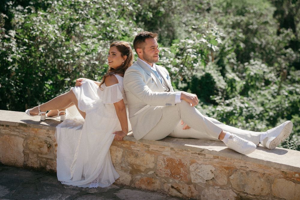 Bride and groom in white wedding attire sitting back-to-back on a stone wall surrounded by lush green trees.