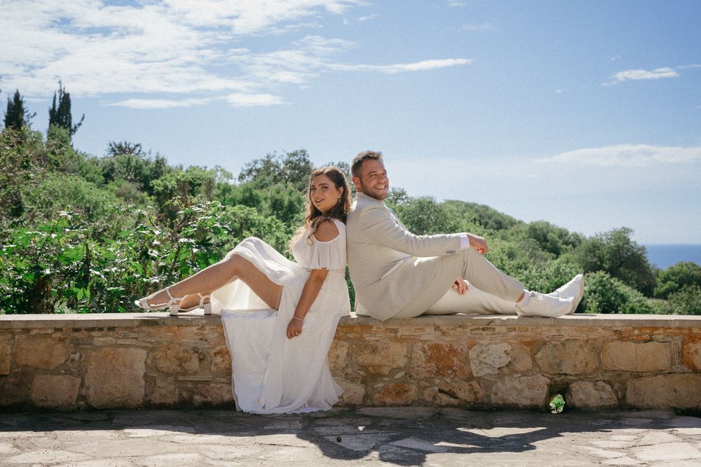 Bride and groom in white attire sitting back-to-back on a stone wall with lush greenery and blue sky behind them.