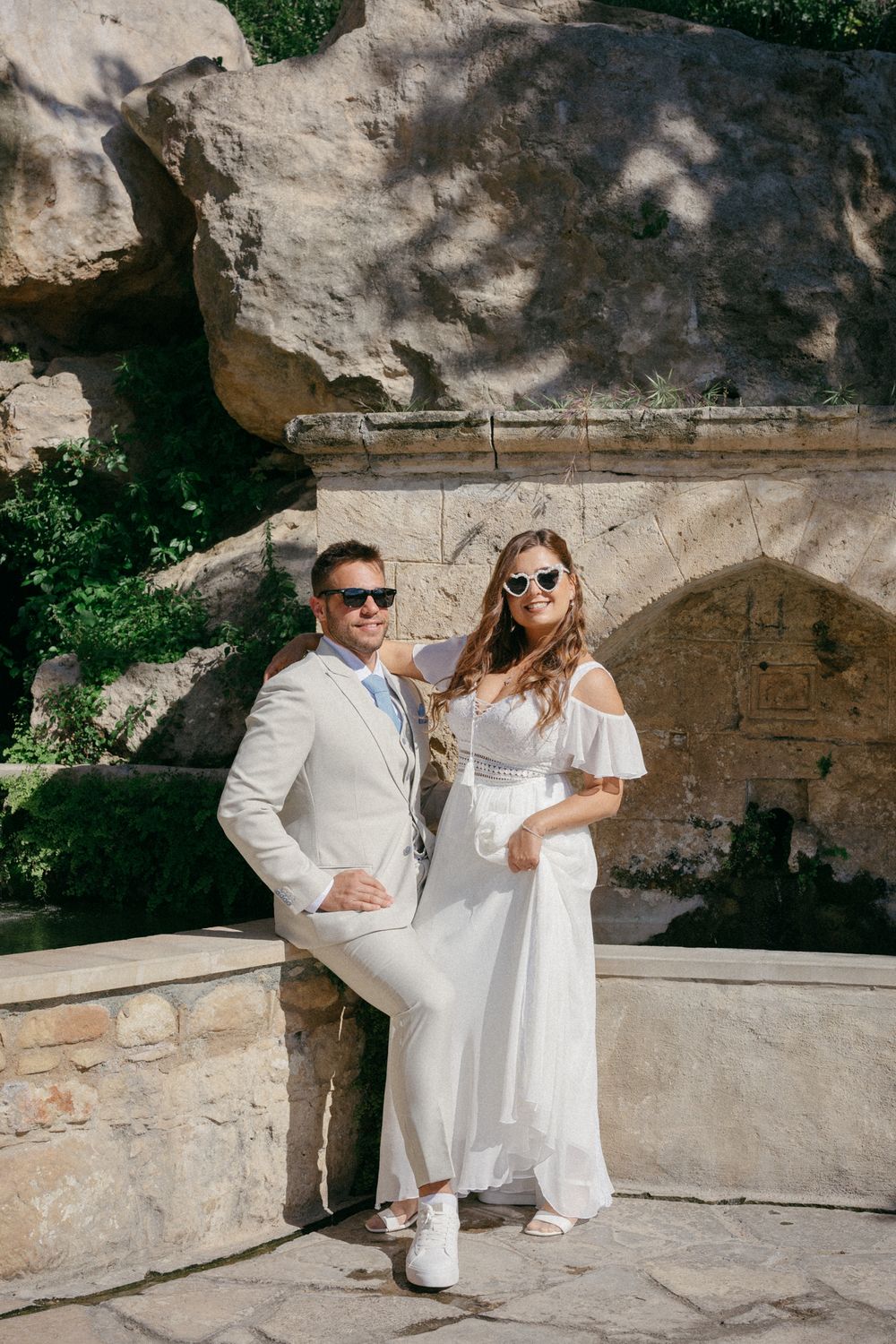 Elegant couple in white wedding attire and sunglasses posing by stone wall in sunny outdoor setting.