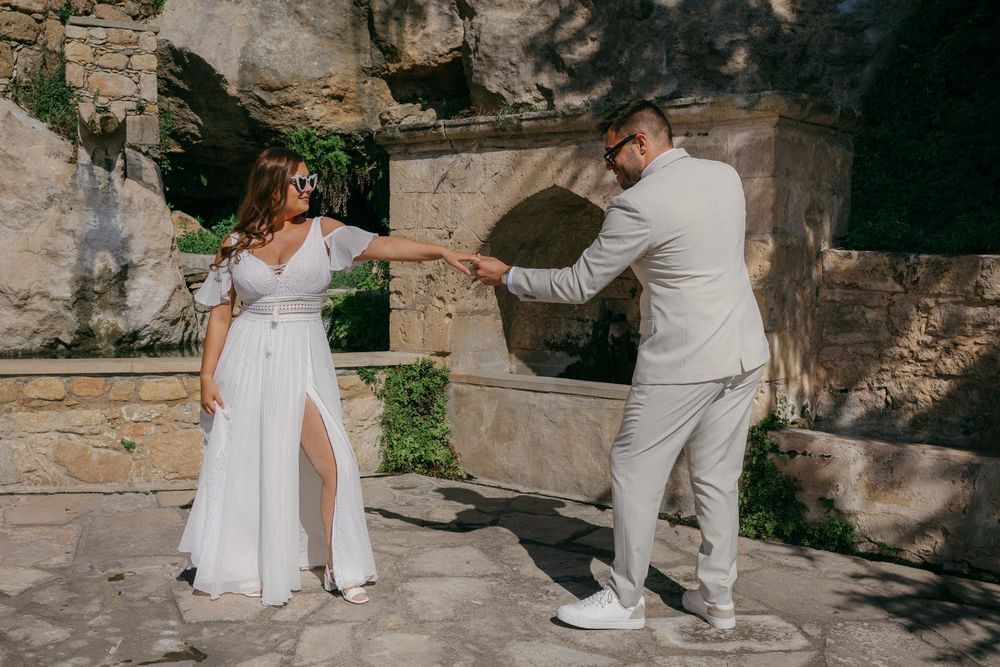 Couple in white wedding attire dancing outdoors near rustic stone wall in sunny courtyard.
