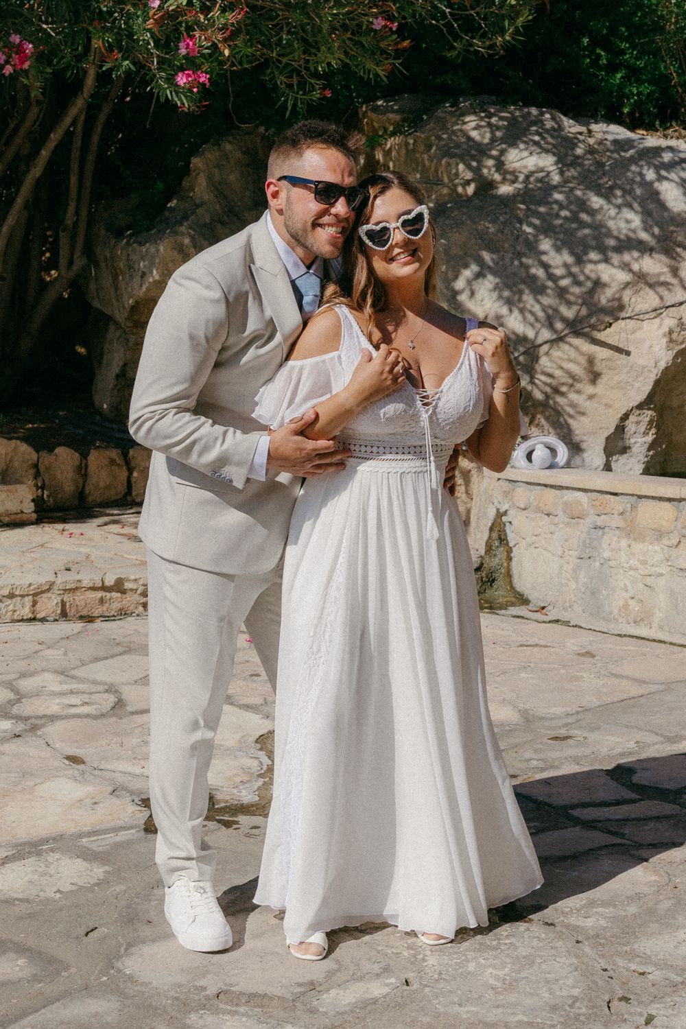 A smiling couple in wedding attire poses outdoors, groom in beige suit, bride in white gown, both wearing stylish sunglasses.