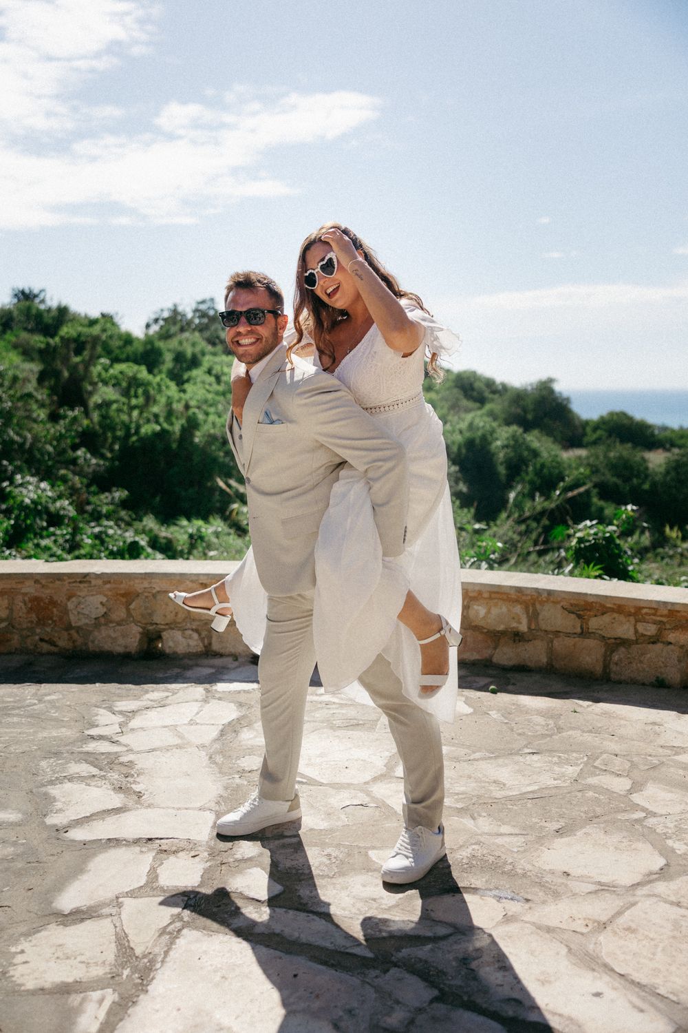 A joyful couple in matching white outfits and sunglasses, with the groom giving the bride a piggyback ride outdoors.