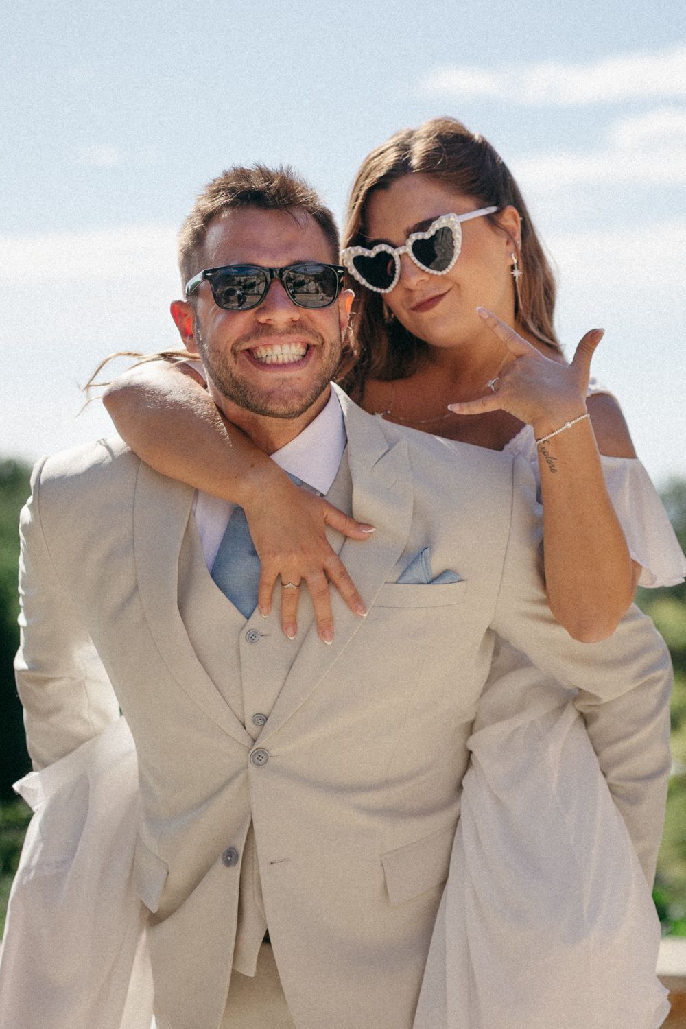 Happy couple in wedding attire, man in beige suit, woman in white dress with heart sunglasses, posing outdoors.