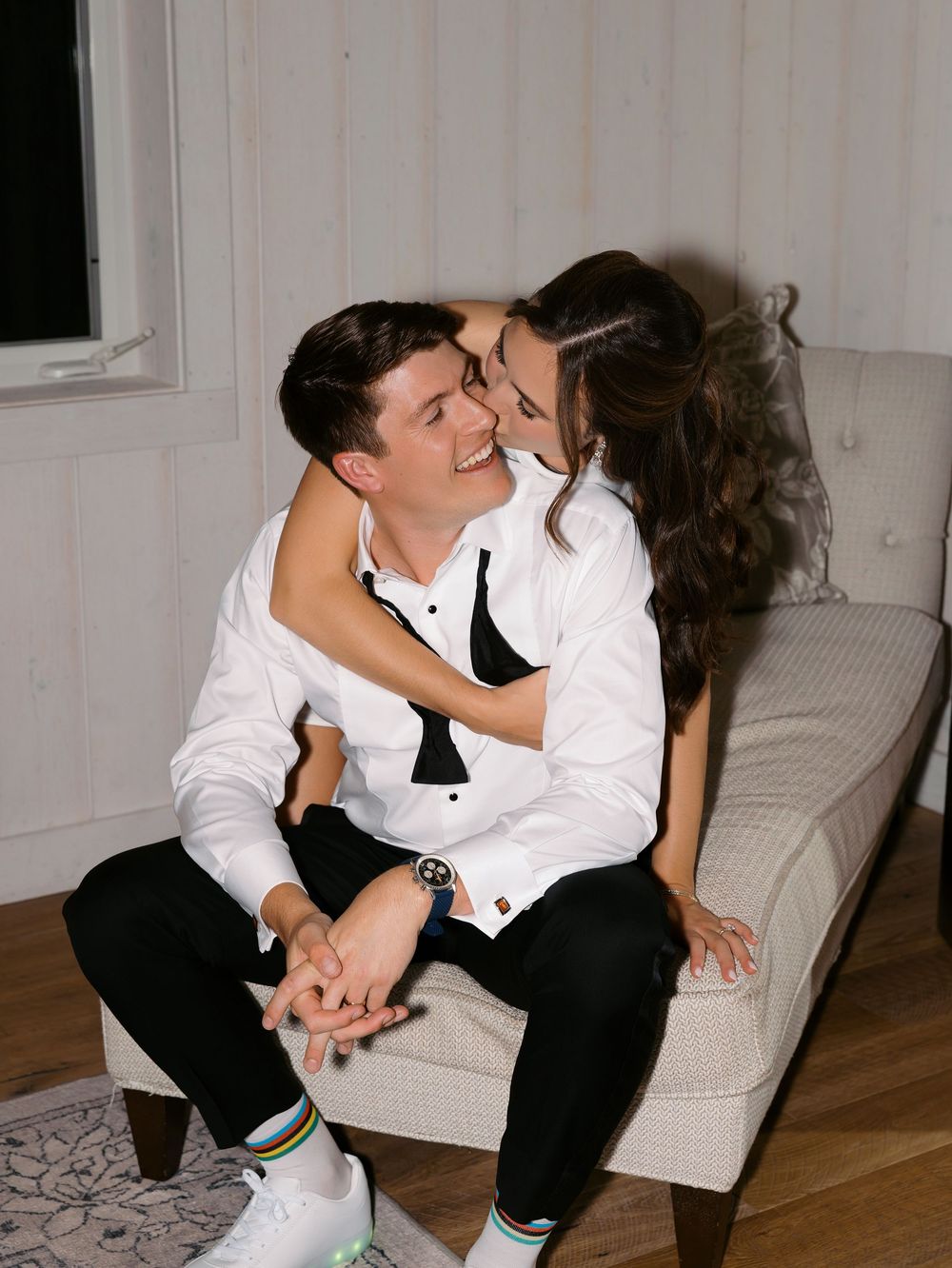 A couple in black and white formal attire share a romantic moment on a chaise lounge, smiling and holding hands.