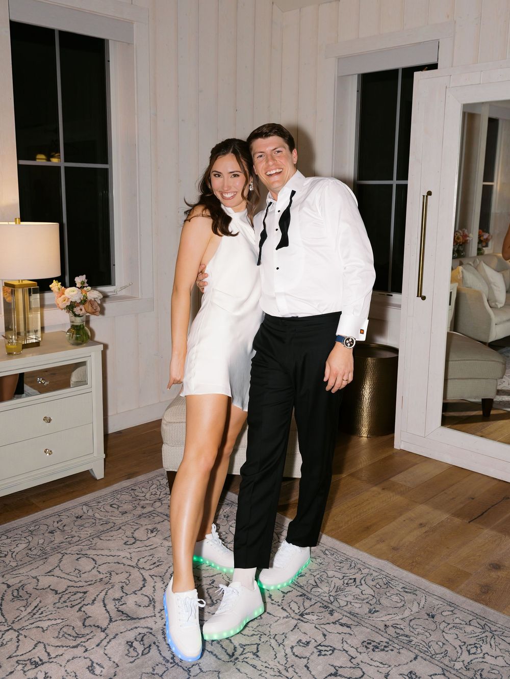 Couple in white dress and tuxedo posing together wearing matching white LED sneakers indoors.