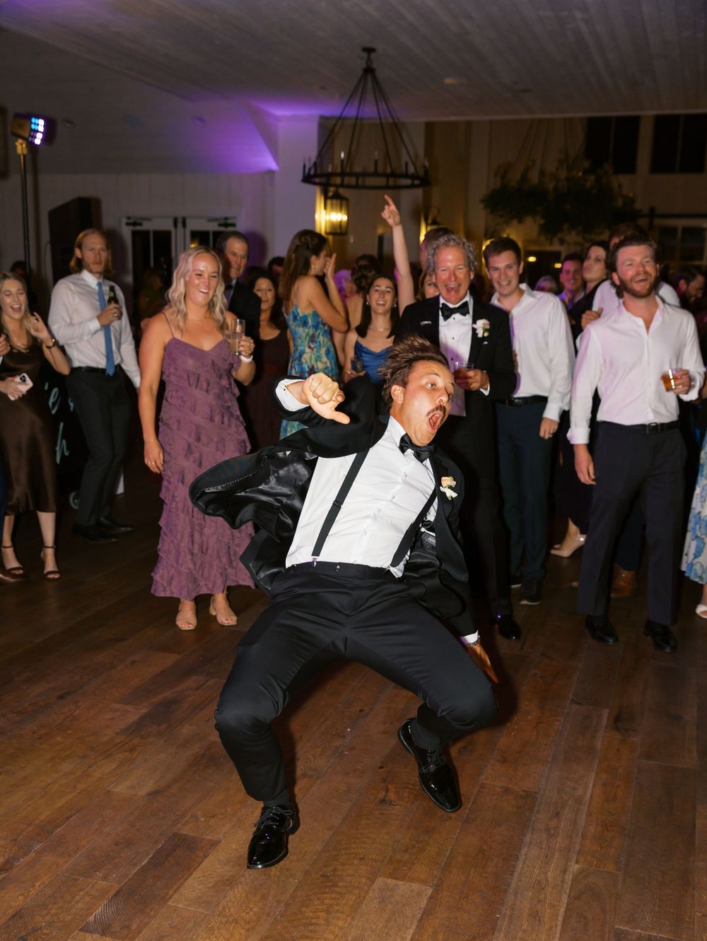 Groom breakdancing at wedding reception while guests cheer on the dance floor.