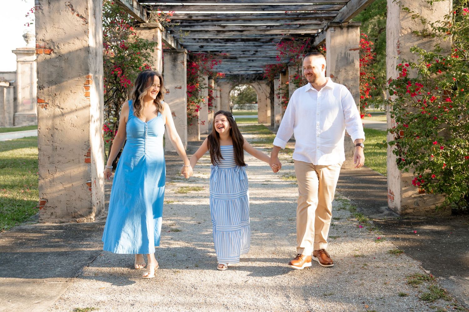 Family of three walks hand-in-hand under flower-covered pergola, wearing coordinated blue and white outfits outdoors.