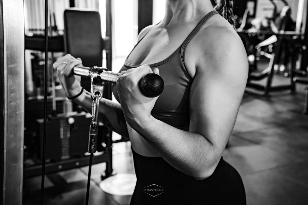 Black and white photo of a muscular woman curling a dumbbell during an intense workout session in a gym.