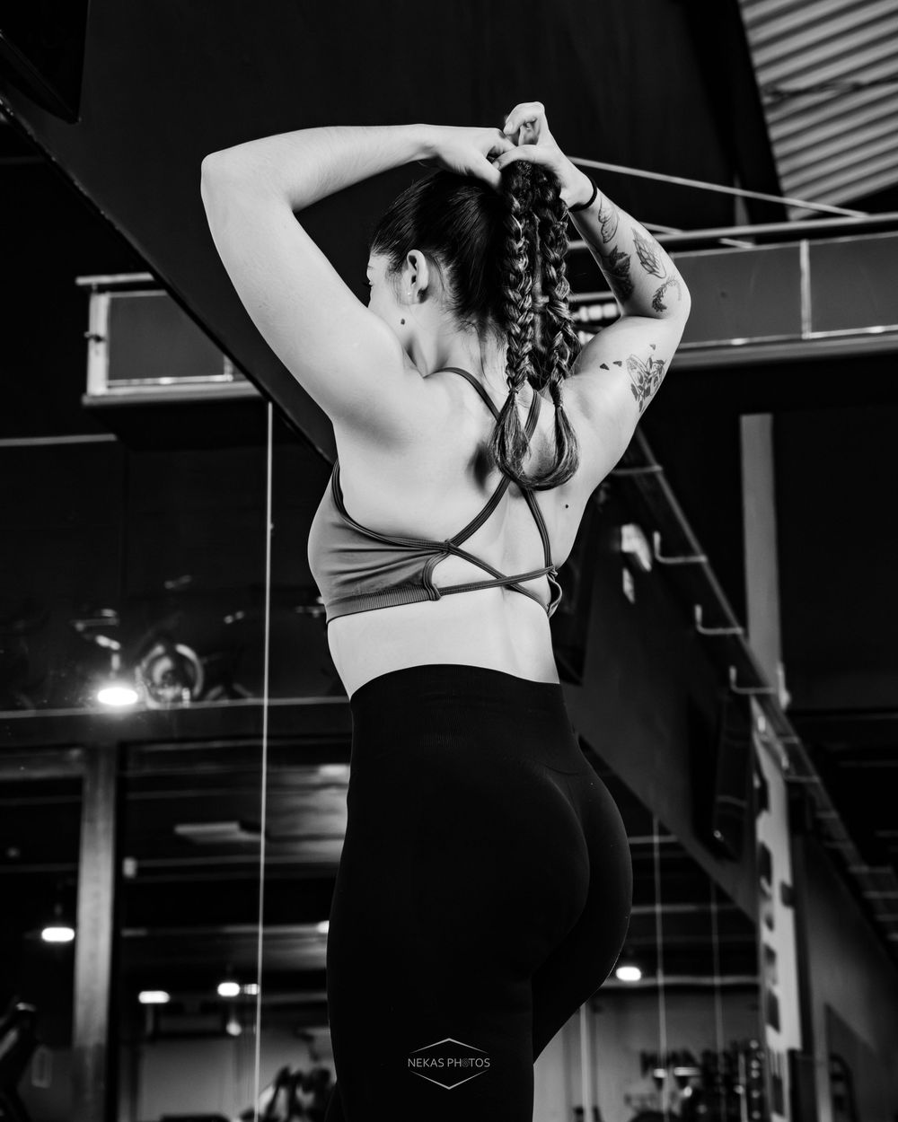 Black and white photo of a fit woman in sports bra and leggings fixing her braided hair in a gym.