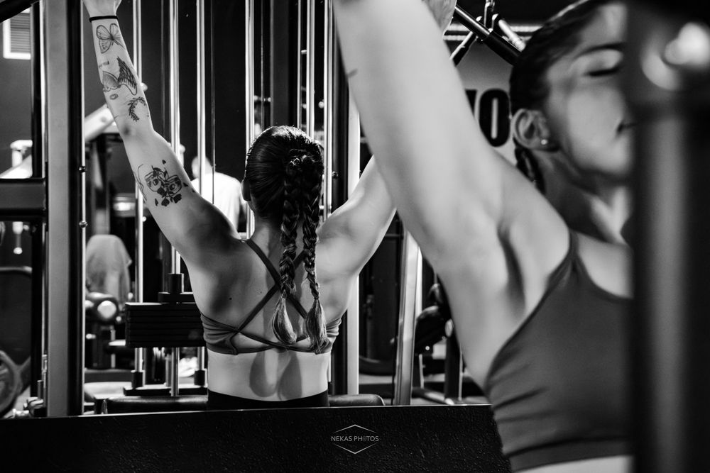 Black and white photo of a woman exercising at a gym, stretching arms overhead near a cable machine.