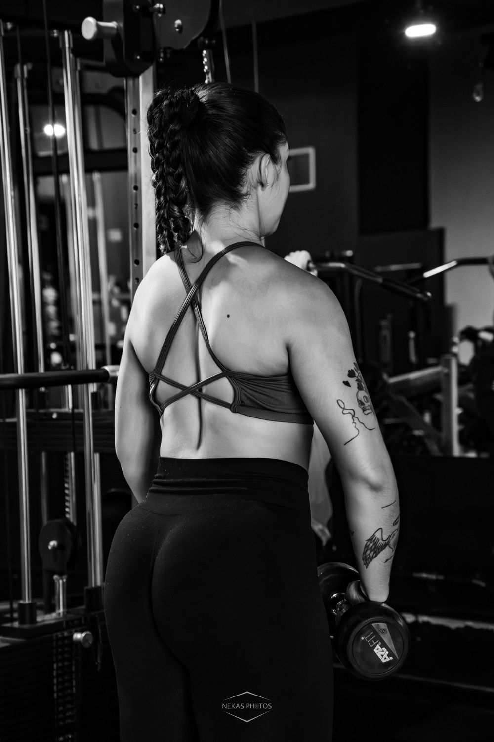 Black and white photo of a fit woman with tattoos lifting dumbbells in a gym, wearing a sports bra and leggings.