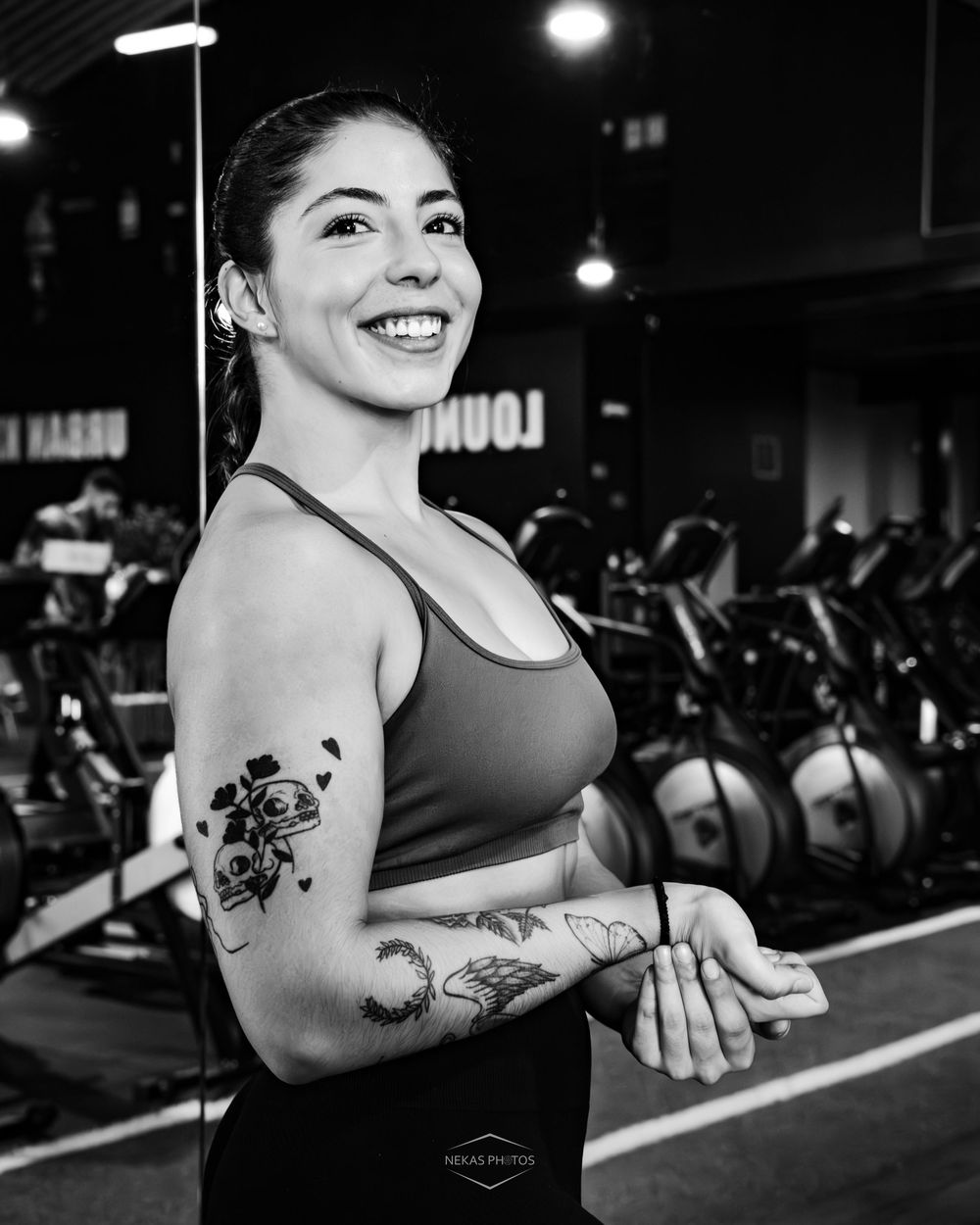 Smiling woman with tattoos in a sports bra poses at a gym, black and white fitness portrait photo.