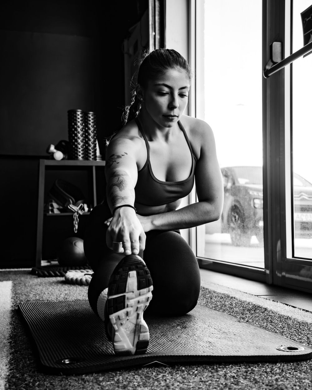 Woman stretching on yoga mat in black and white photo, reaching toward sneaker in home gym near window.