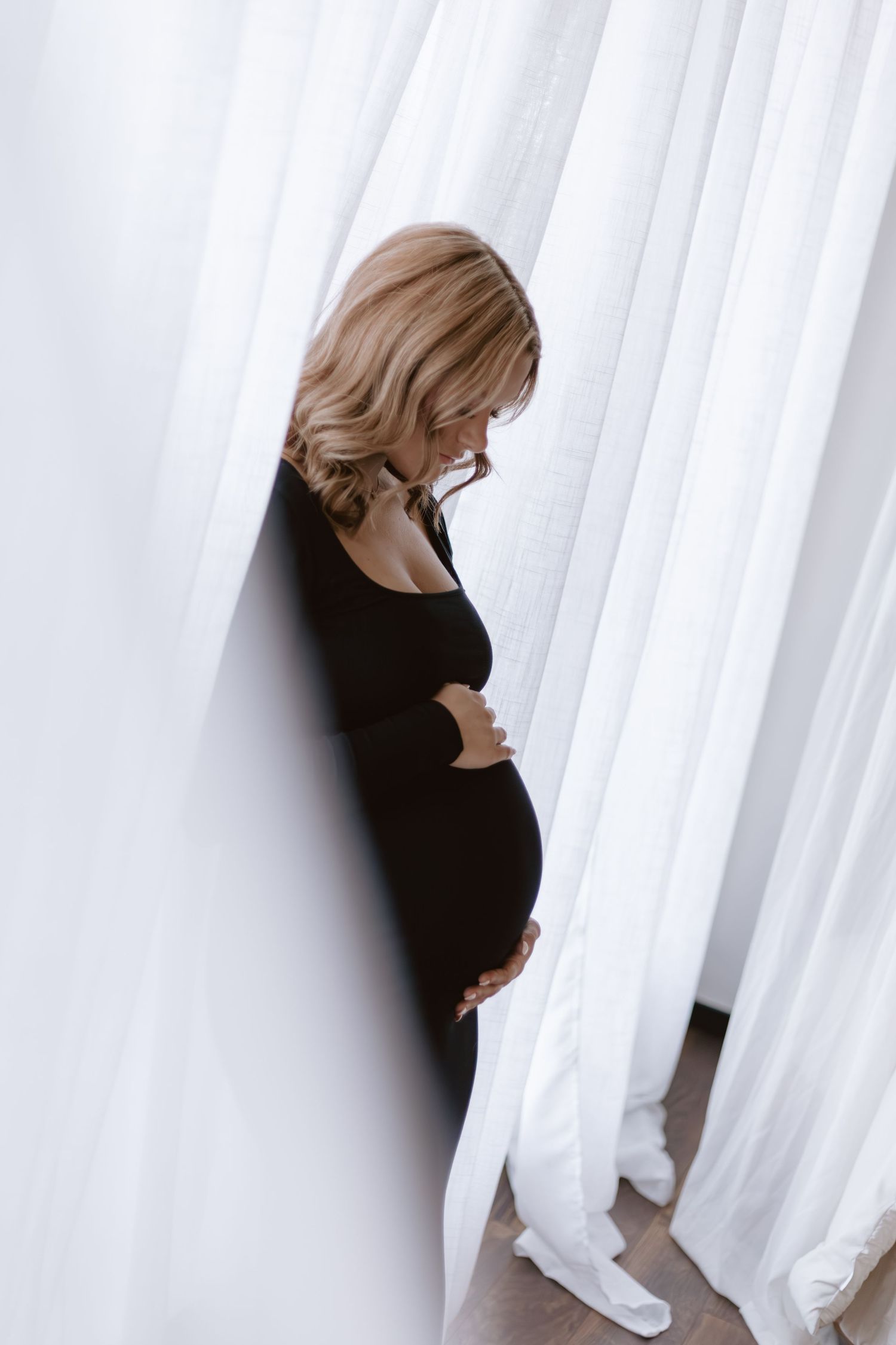 Pregnant woman in black dress standing between white curtains gently cradling her belly.