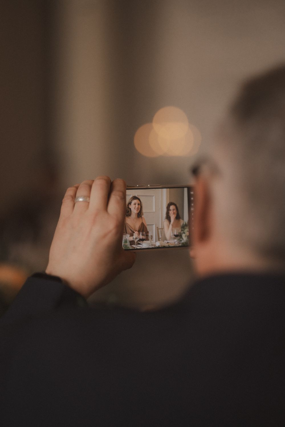 Person holding smartphone horizontally capturing photo of two smiling women at dinner table, wearing wedding ring.