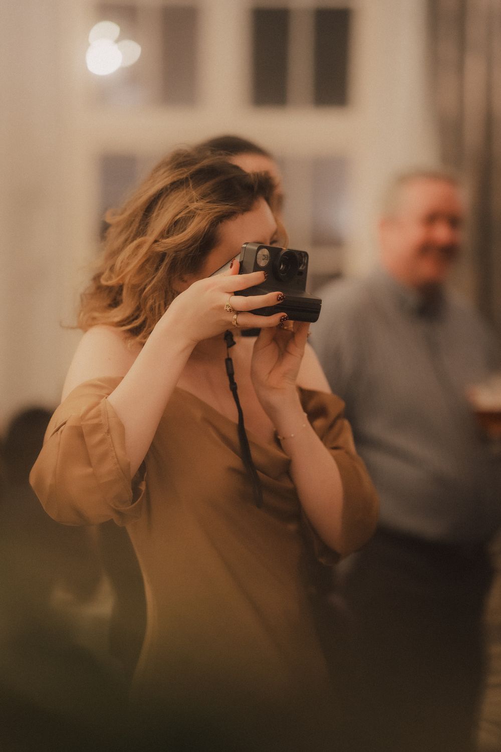 Woman with curly hair photographing with black camera at indoor social gathering in warm light.