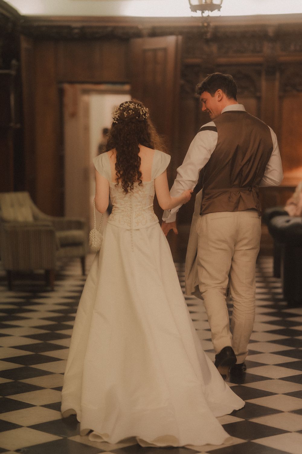 Bride in white lace gown and groom in vest hold hands walking across checkered floor in elegant wood-paneled venue.