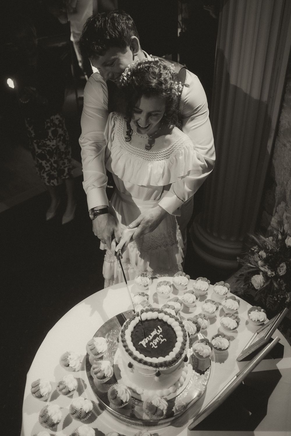 Bride and groom cutting wedding cake together at reception, black and white photo with cupcakes displayed nearby.