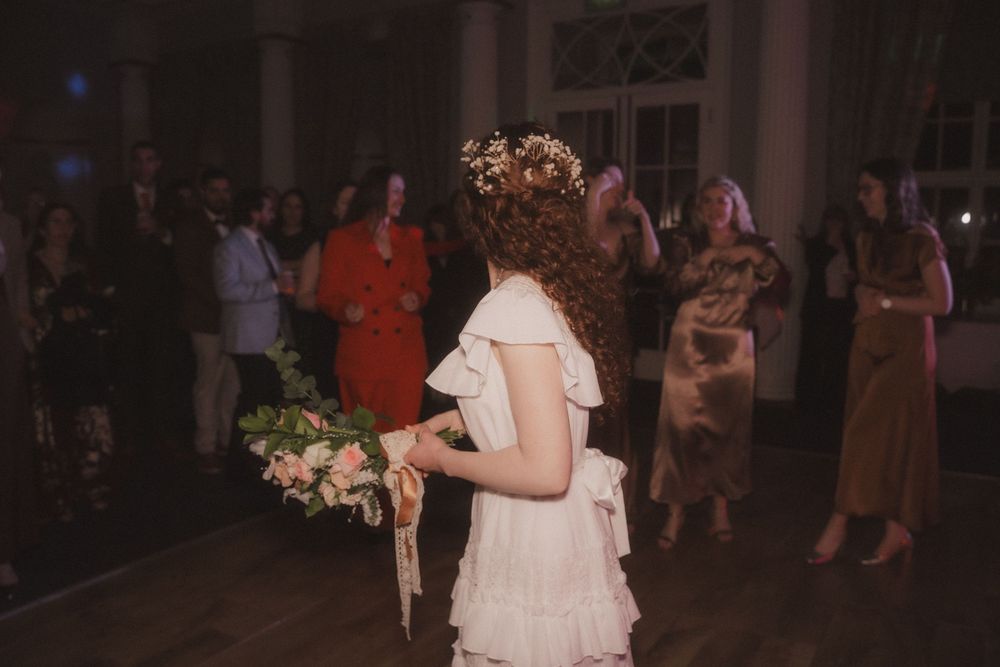 Bride in white ruffled dress holding floral bouquet with flower crown, surrounded by guests at elegant wedding reception.