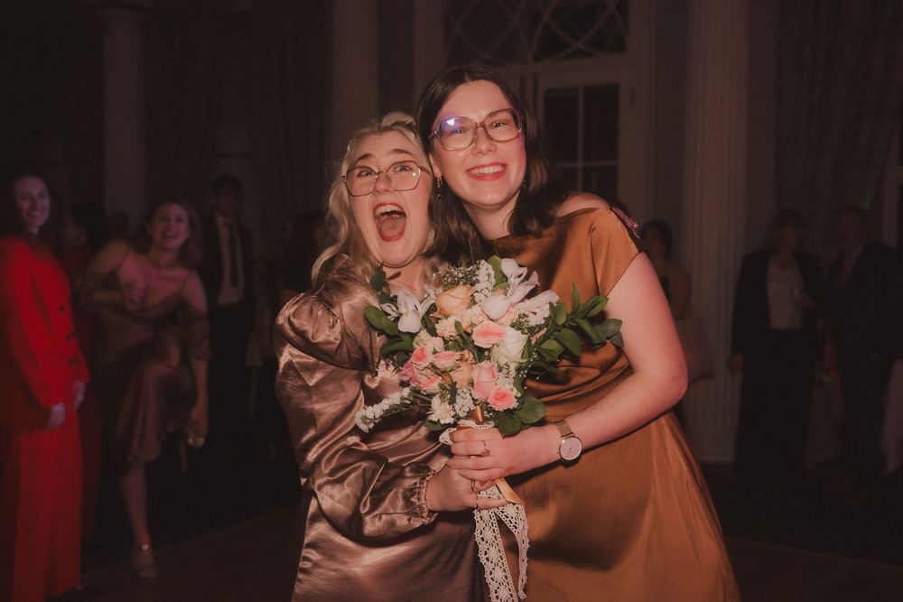 Two women in gold satin dresses smiling and hugging while holding a pink and white bouquet at a wedding reception.