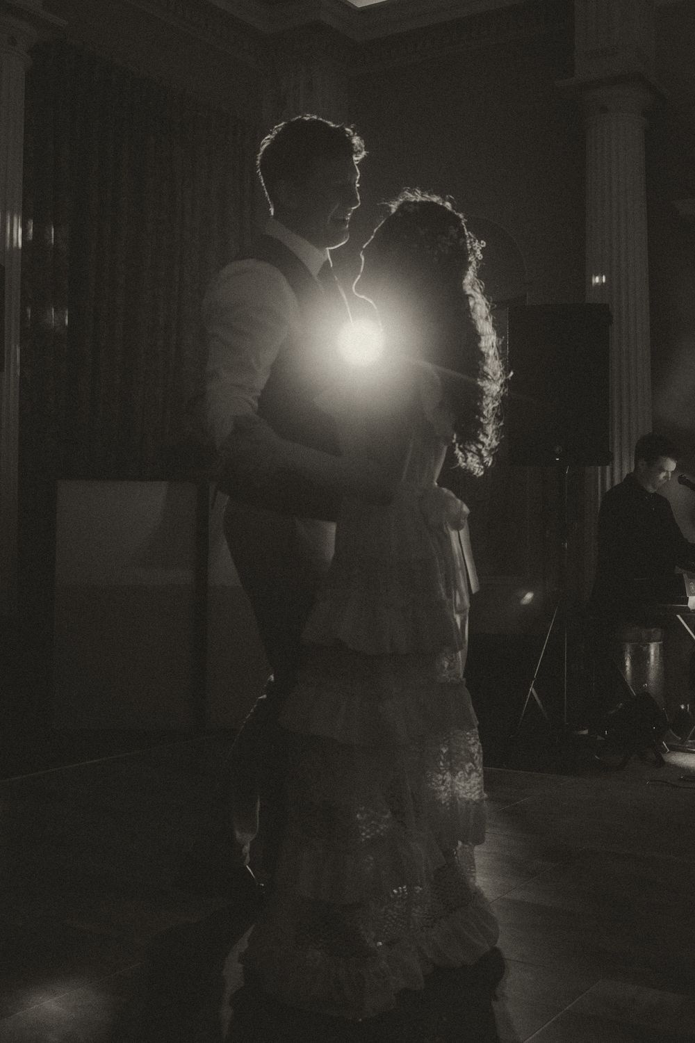 Black and white silhouette of a couple sharing first dance at wedding reception with dramatic backlight glow.