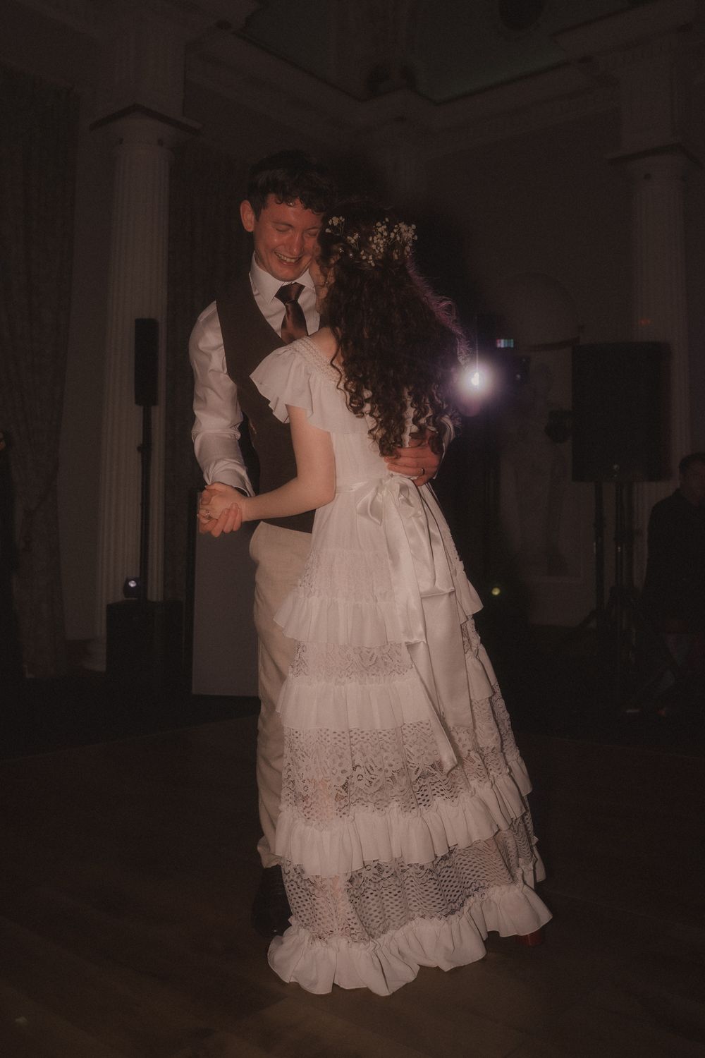 Bride and groom share their first dance in a dimly lit elegant hall, bride wearing a tiered lace gown with floral hair.