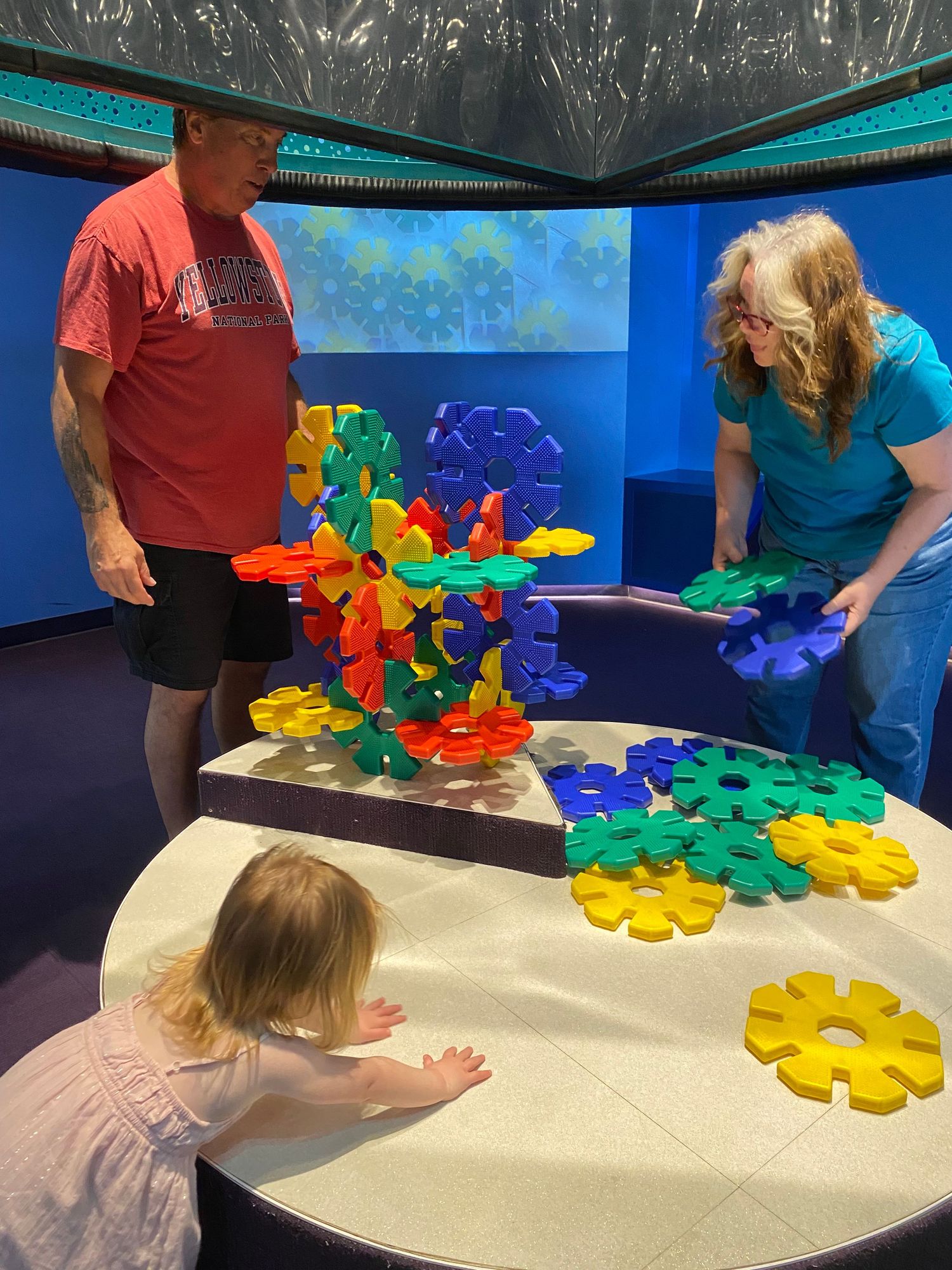 Two adults and a toddler play with colorful interlocking gear toys at a children's museum interactive exhibit.